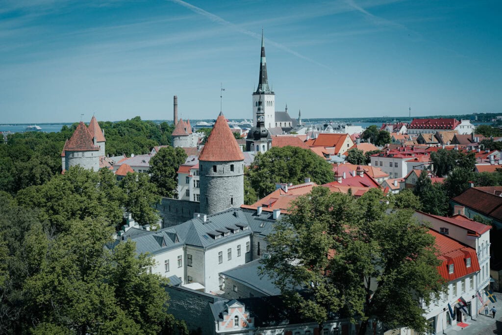 Eine Stadtlandschaft mit Gebäuden mit roten Dächern, Steintürmen und einem hohen Kirchturm zwischen grünen Bäumen unter einem blauen Himmel.