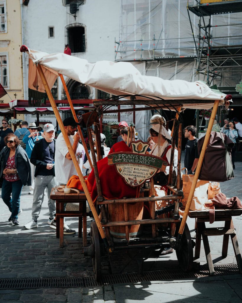 Auf einem mittelalterlich angehauchten Straßenkarren in Tallinn sind kostümierte Menschen zu sehen, die auf einem belebten Stadtplatz von Passanten umgeben sind.