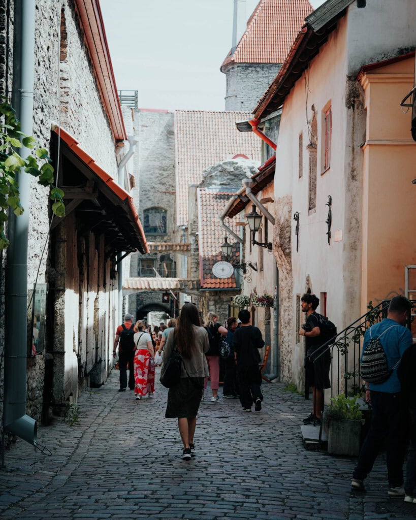 Menschen schlendern durch eine schmale Kopfsteinpflasterstraße in Tallinn, gesäumt von alten Gebäuden aus Stein und Stuck, und genießen den sonnigen Tag.
