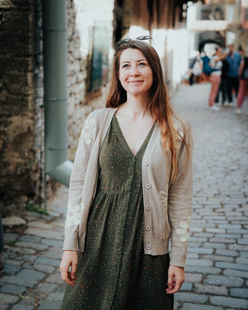 Eine Frau in einem grünen Kleid und einer beigen Strickjacke steht lächelnd auf einer Kopfsteinpflasterstraße in Tallinn.