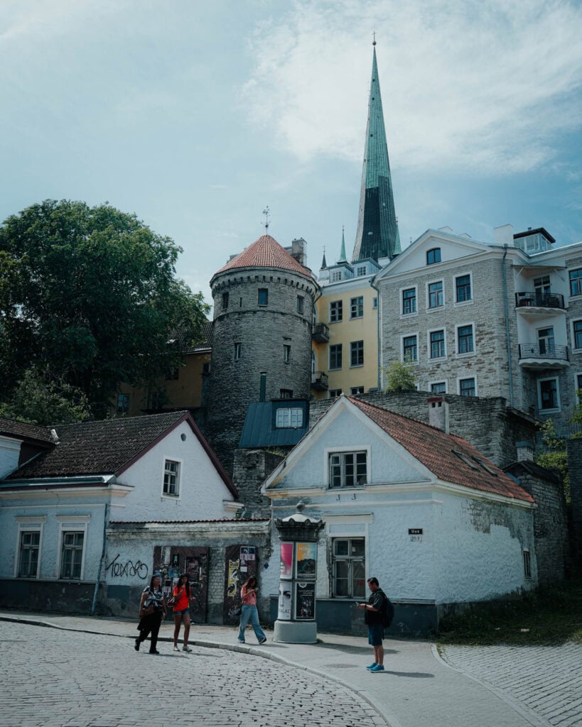 Unter einem teilweise bewölkten Himmel gehen Menschen in Tallinn an alten Steingebäuden und einem Turm mit spitzer Spitze vorbei.