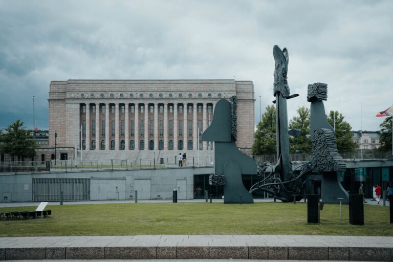 Moderne abstrakte Skulpturen auf einer Wiese mit einem großen neoklassizistischen Gebäude im Hintergrund unter bewölktem Himmel.
