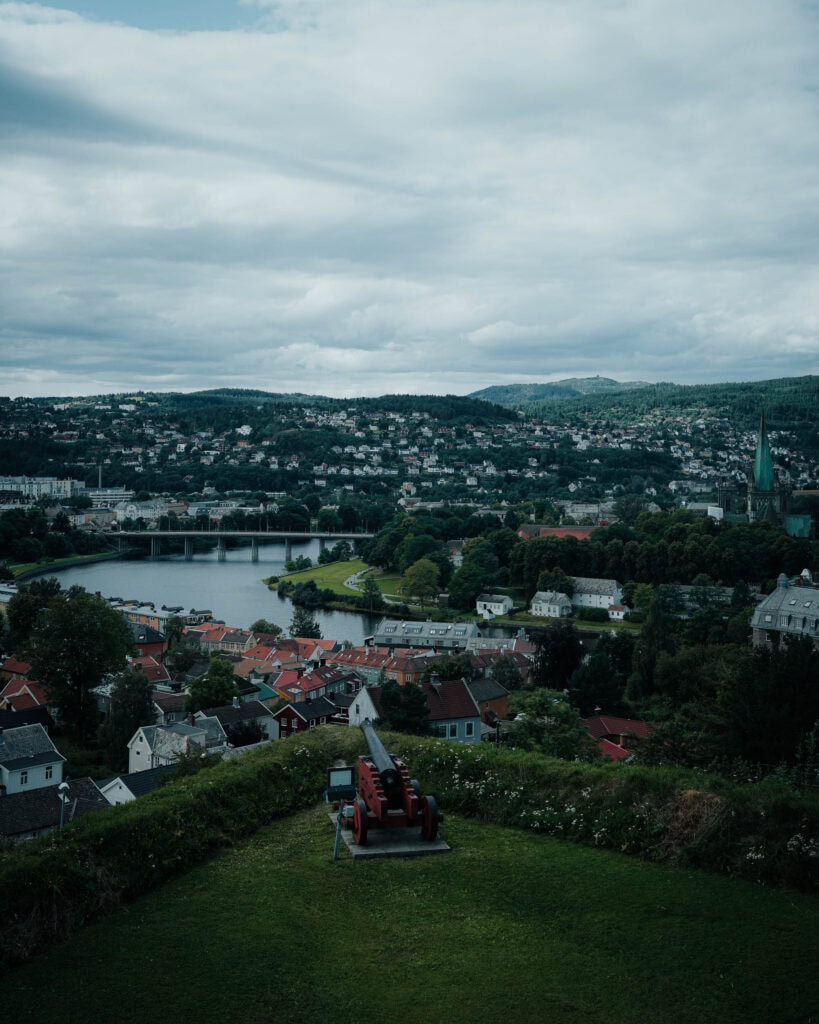 Blick von einer Hügelkuppe mit einer roten Kanone auf eine Stadt, einen Fluss und ferne grüne Hügel bei bewölktem Himmel.