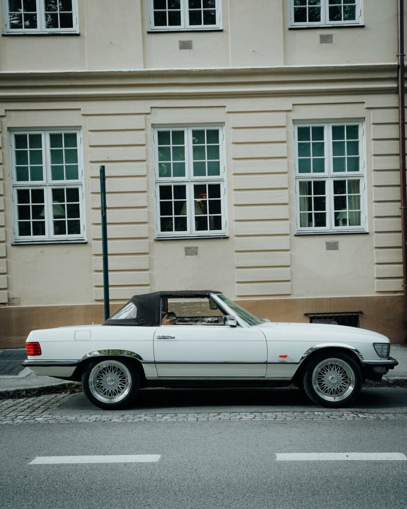 Ein klassisches weißes Cabrio, das vor einem beigen Gebäude mit großen Fenstern geparkt ist.