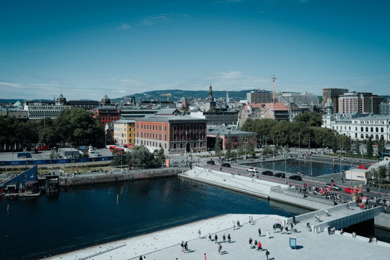 Eine Stadtlandschaft mit bunten Gebäuden am Wasser, Menschen, die unter blauem Himmel am Wasser spazieren gehen.
