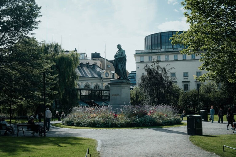Statue in der Mitte eines Parks, umgeben von Blumen, Bäumen und Gebäuden an einem sonnigen Tag.
