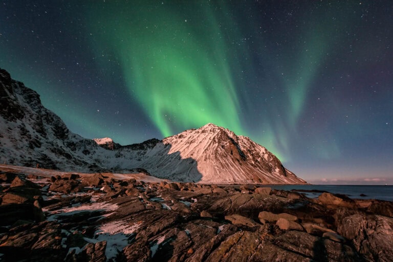 Das Polarlicht leuchtet grün über schneebedeckten Bergen und felsigen Ufern unter einem sternenklaren Nachthimmel.
