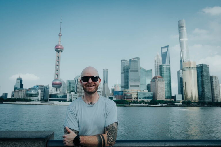 Ein lächelnder Mann mit Tattoos und Sonnenbrille steht an einem Fluss, mit der Skyline von Shanghai im Hintergrund.