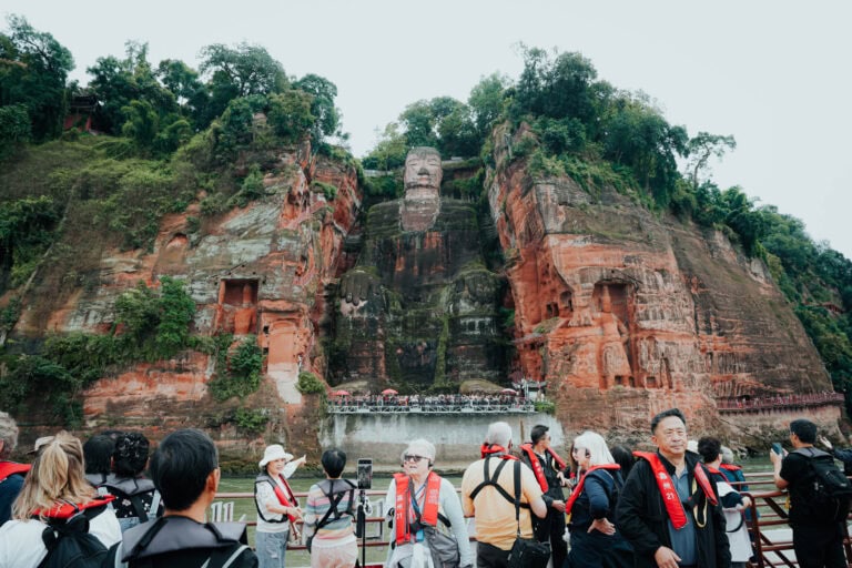 Eine Gruppe von Menschen steht vor einer großen Felswand mit dem Riesenbuddha von Leshan im Hintergrund.