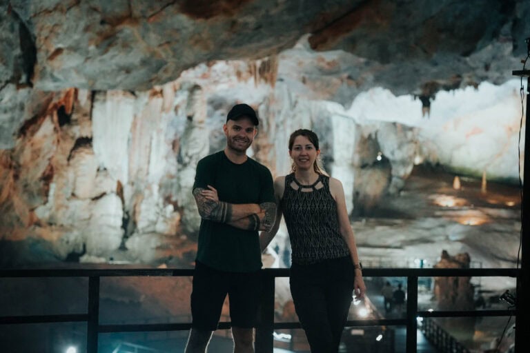 Zwei Personen stehen lächelnd in einer großen, beleuchteten Höhle mit Felswänden und Felsformationen im Hintergrund.
