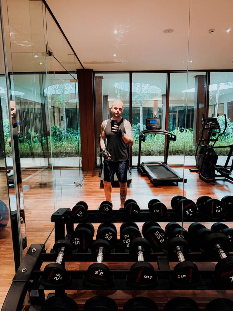 Ein Mann macht ein Selfie in einem Fitnessstudio mit Hanteln, Laufband und Trainingsgeräten, die sich in Spiegeln spiegeln.