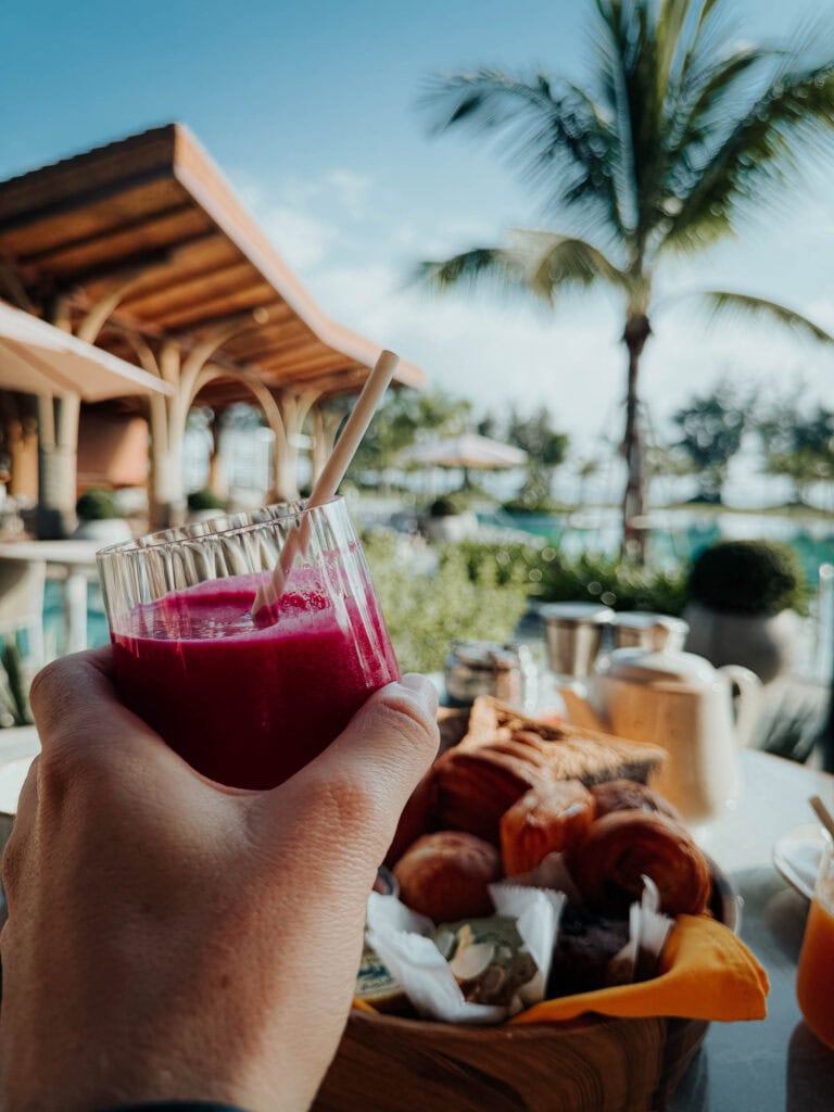 Eine Hand hält ein Glas roten Saft bei einem Frühstück am Pool mit Gebäck, Palme und Resort im Hintergrund.