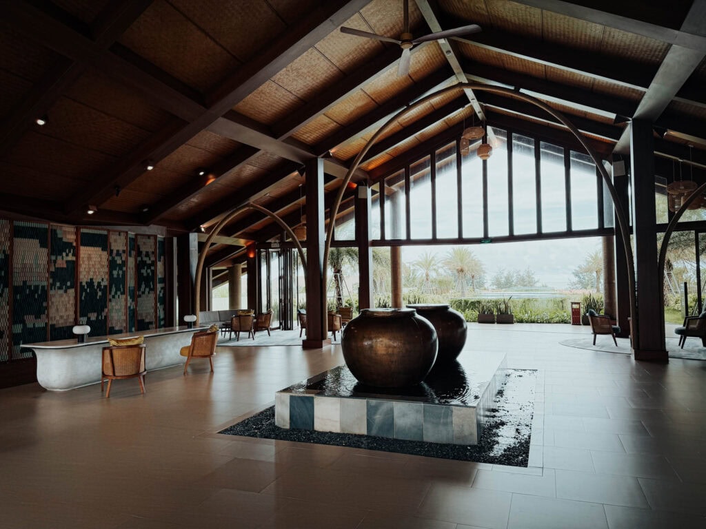 Moderne Hotellobby mit großem Wasserspiel, hohen Holzdecken und Gartenblick durch Glasfenster.