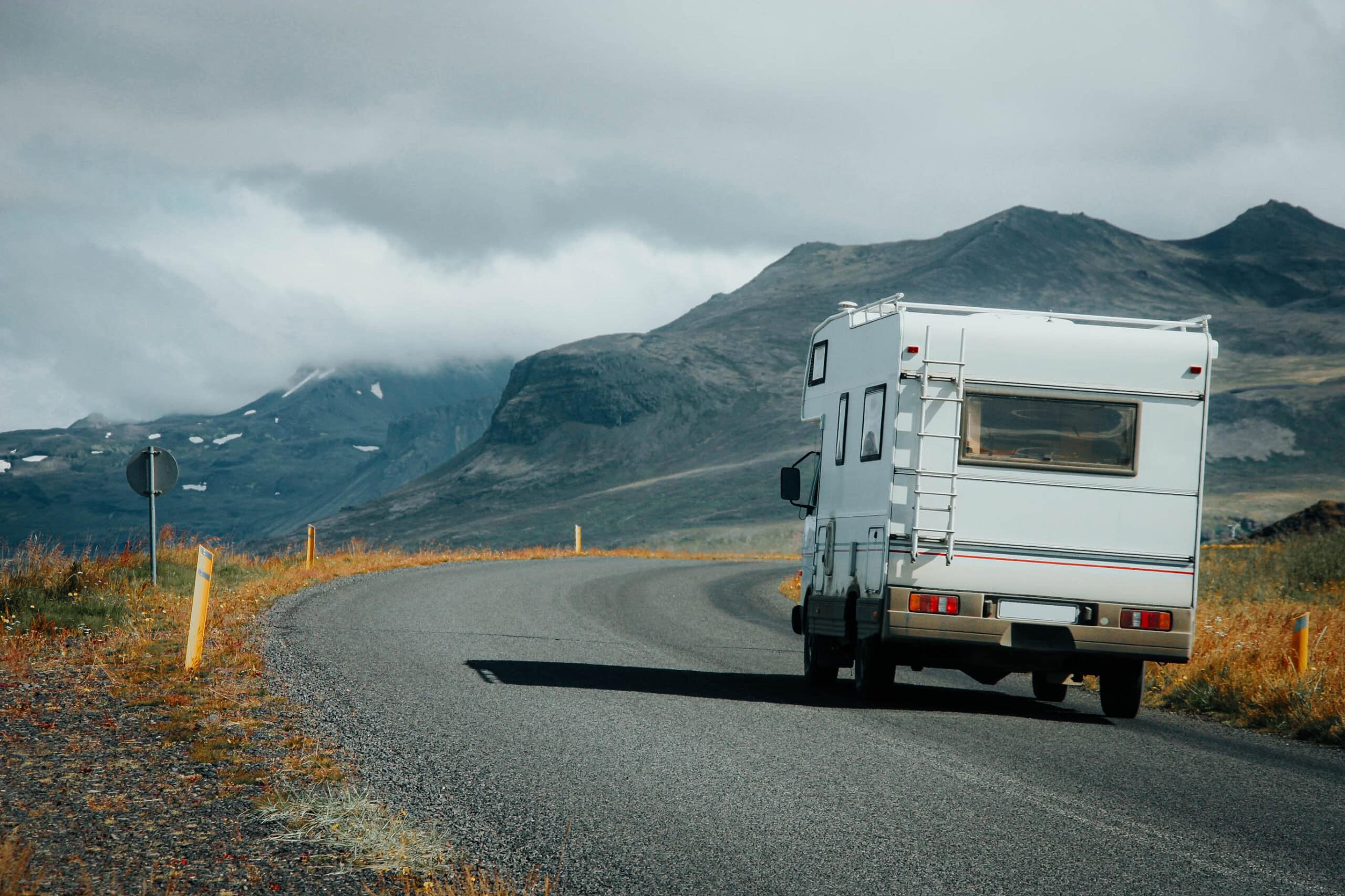 Ein weißes Wohnmobil fährt auf einer kurvenreichen Straße mit Bergen und Wolken im Hintergrund.