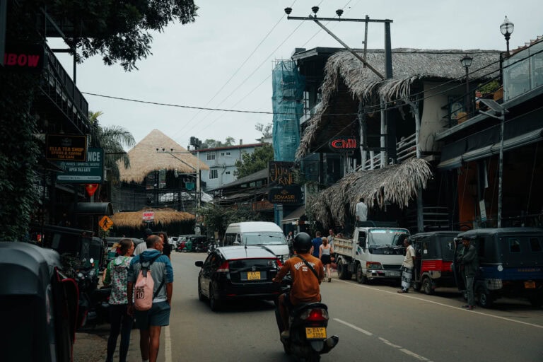 Belebte Straßenszene mit Autos, Motorrädern, Fußgängern, Geschäften und Cafés mit Strohdächern und Schildern.