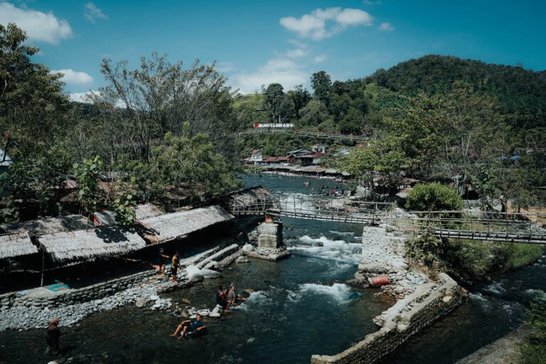 Menschen, die in einem Fluss neben Holzhütten und einem Steg schwimmen, mit üppig grünen Hügeln im Hintergrund.