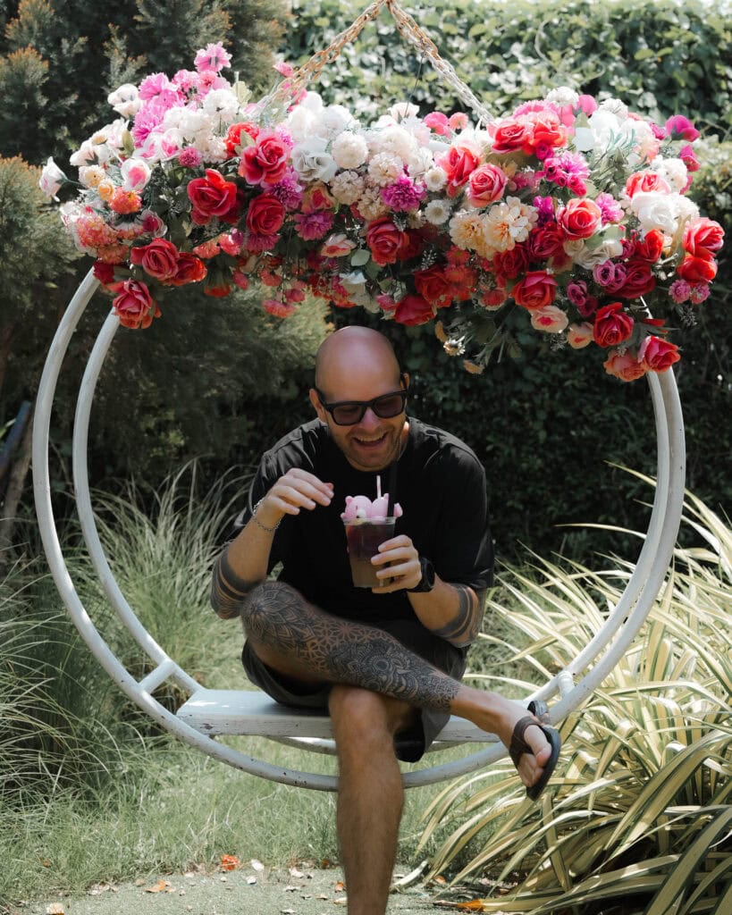 Mann mit Glatze und Brille sitzt auf einer Blumenschaukel im Freien, lächelt und hält ein Getränk mit einem rosa Strohhalm in der Hand.