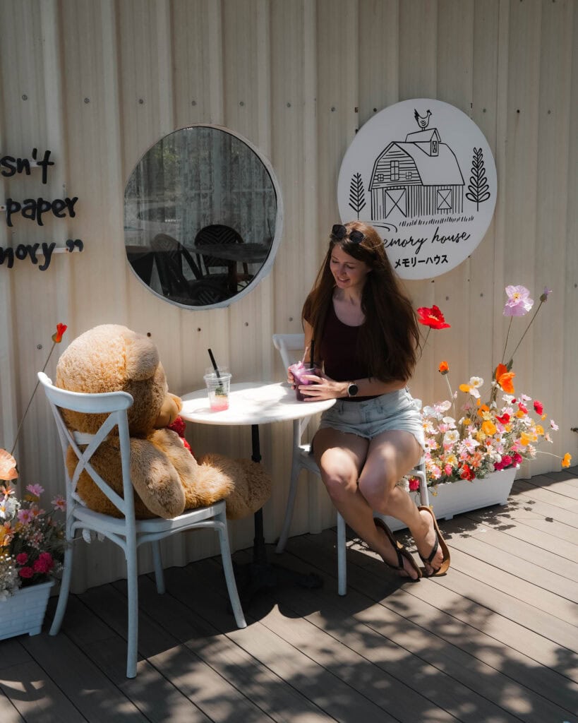 Frau mit Sonnenbrille sitzt an einem kleinen Tisch im Freien mit einem großen Teddybären, Getränken und Blumen in der Nähe.