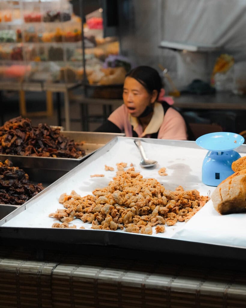 Tablett mit frittierten Snacks mit blauer Skala an einem Marktstand, Verkäufer sitzt hinter der Theke.