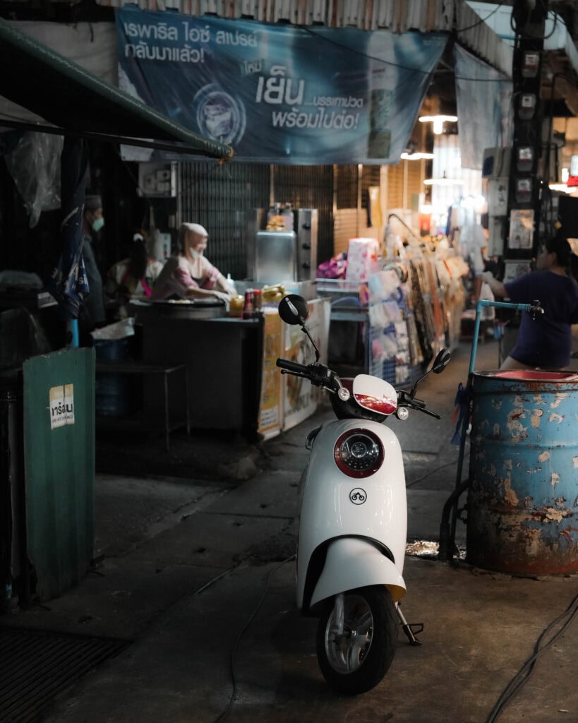 Ein weißer Motorroller, der auf einem schwach beleuchteten Markt mit Essensständen und Verkäufern im Hintergrund geparkt ist.