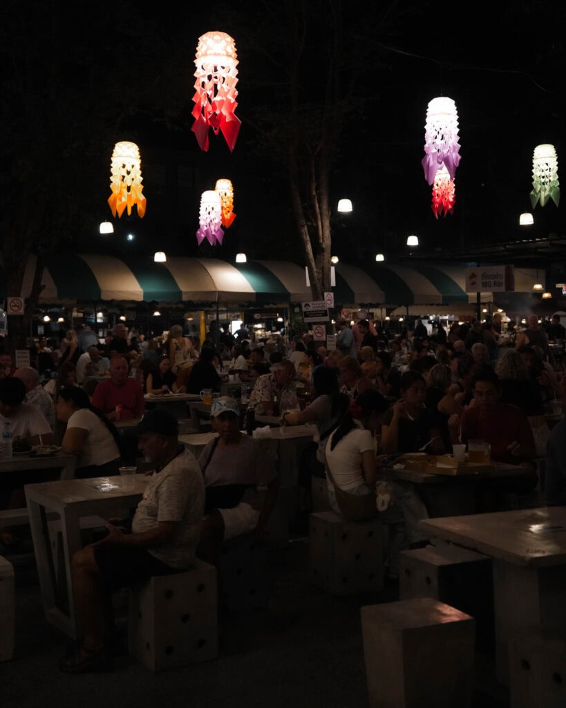Nachtmarkt im Freien mit Menschen, die an Tischen unter bunten hängenden Laternen speisen.