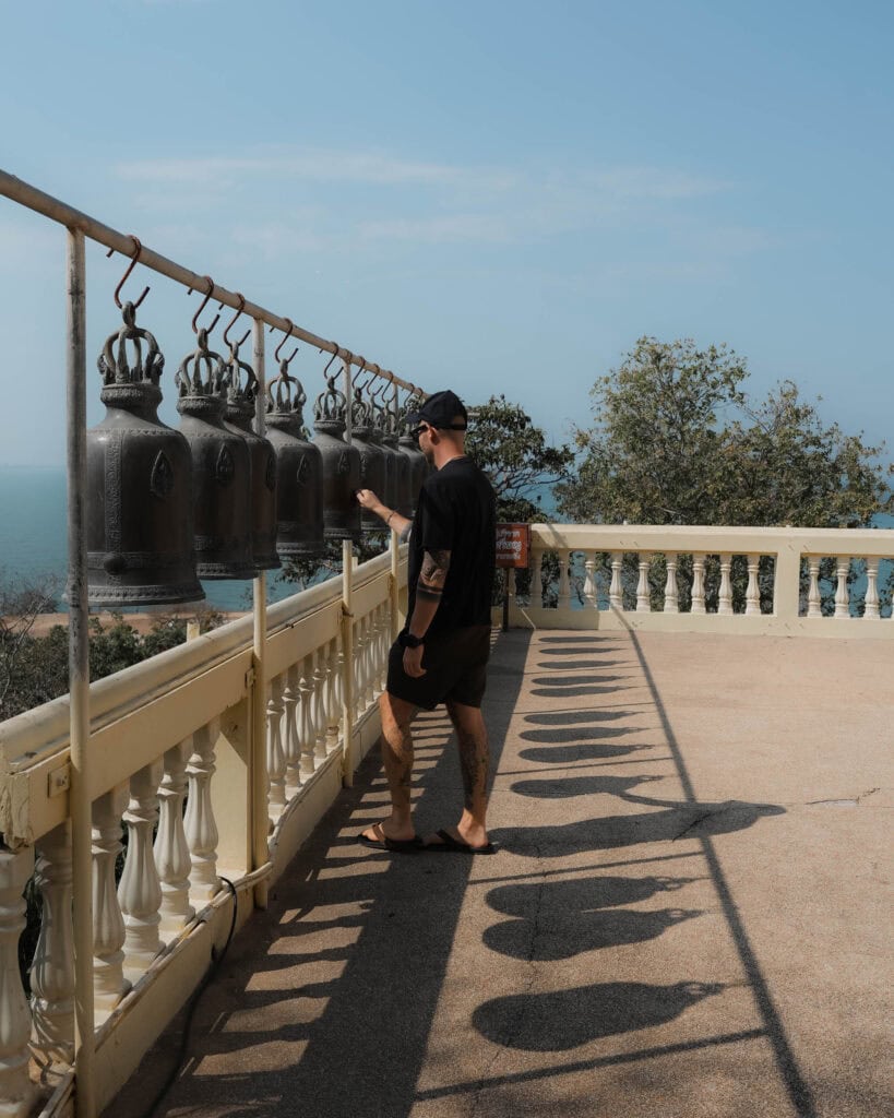 Eine Person läutet große hängende Glocken auf einer sonnigen Terrasse mit Blick aufs Meer.