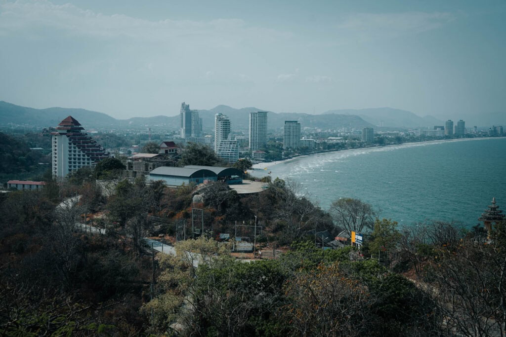 Eine Stadtlandschaft an der Küste mit hohen Gebäuden, einer geschwungenen Küstenlinie und Bergen im Hintergrund.