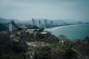 Eine Stadtlandschaft an der Küste mit hohen Gebäuden, einer geschwungenen Küstenlinie und Bergen im Hintergrund.