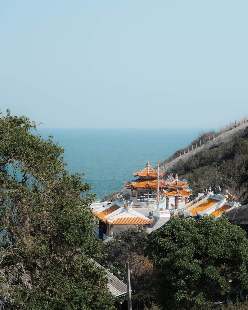 Ein traditioneller Tempel mit orangefarbenen Dächern blickt auf das blaue Meer, umgeben von Grün und Hügeln.