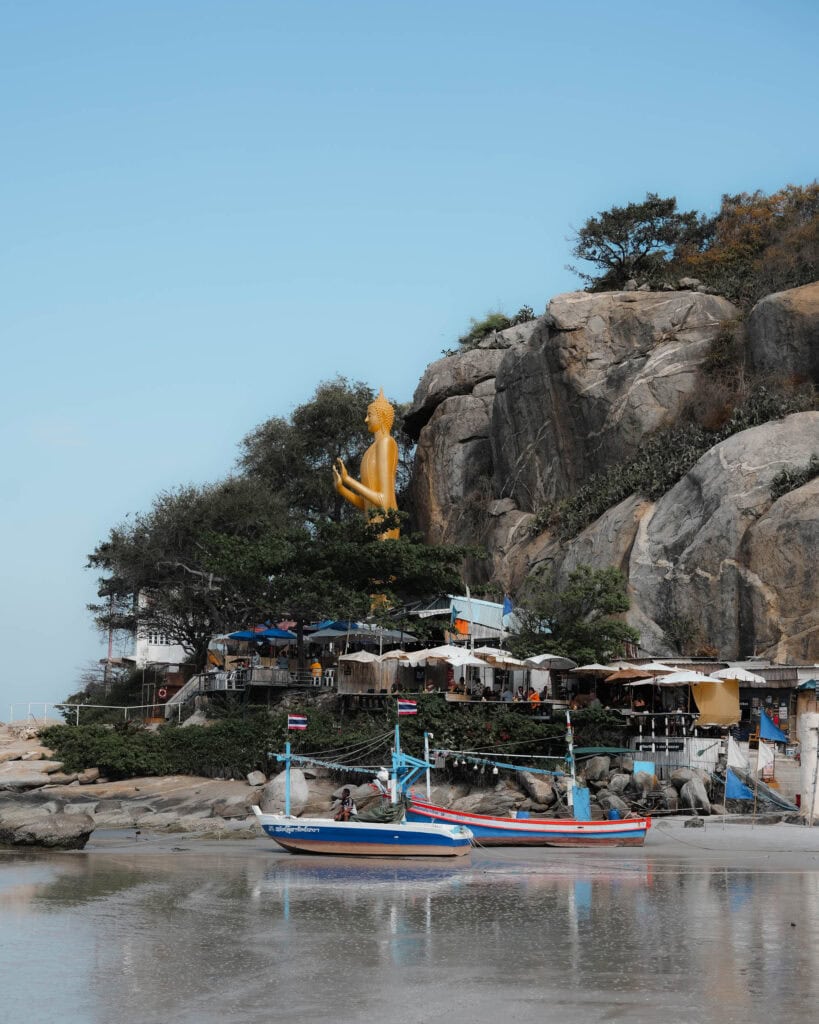 Eine große goldene Buddha-Statue überblickt bunte Boote an einem felsigen Abhang und Gebäude am Wasser.