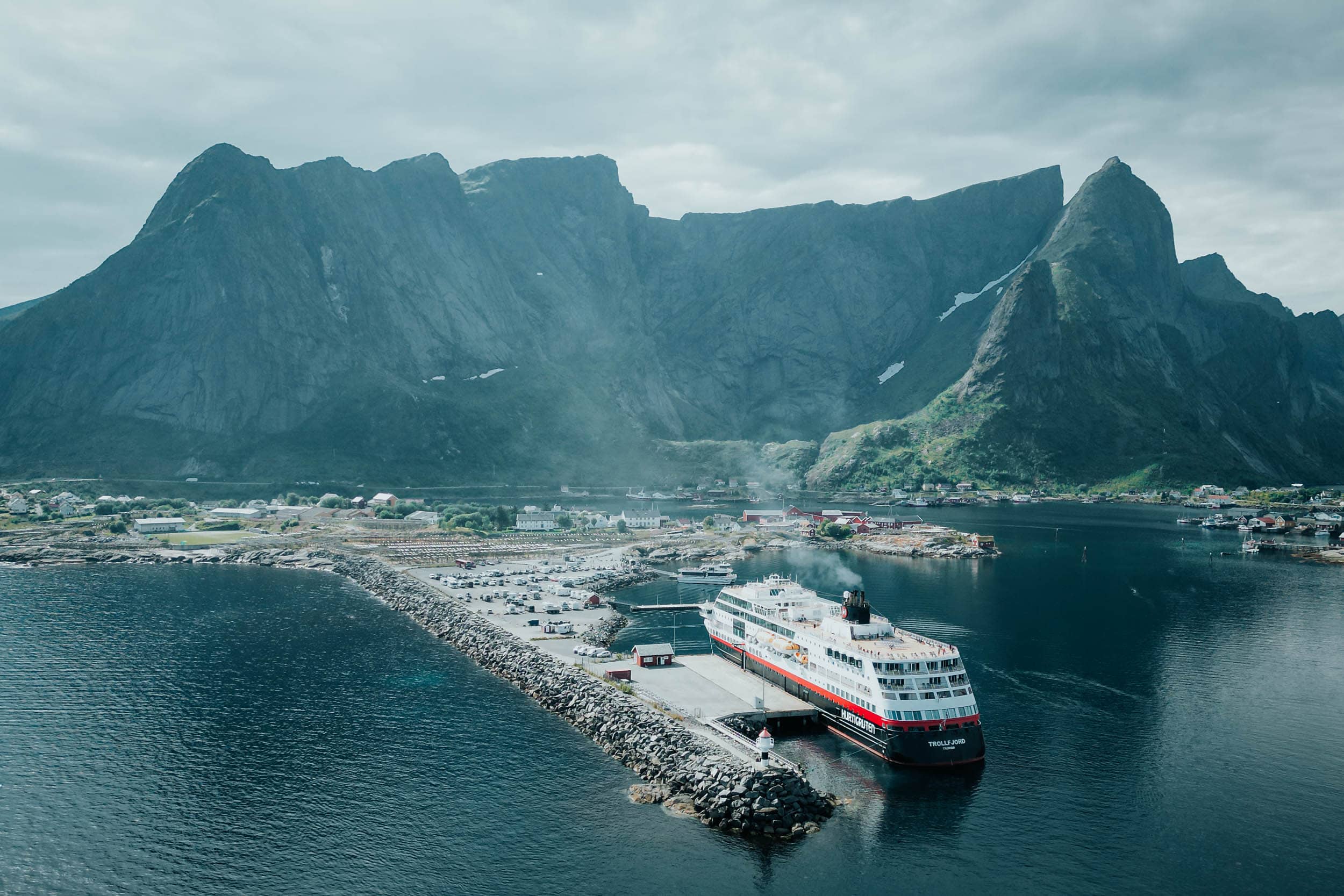 Eine große Fähre, die an einer Anlegestelle anlegt, mit Bergen und einem kleinen Küstendorf im Hintergrund.
