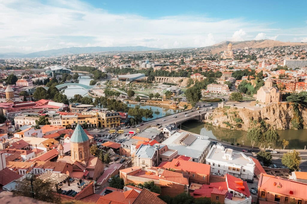 Stadtbild von Tiflis, Georgien, mit Gebäuden mit roten Dächern, Fluss, Brücken und Hügeln im Hintergrund.