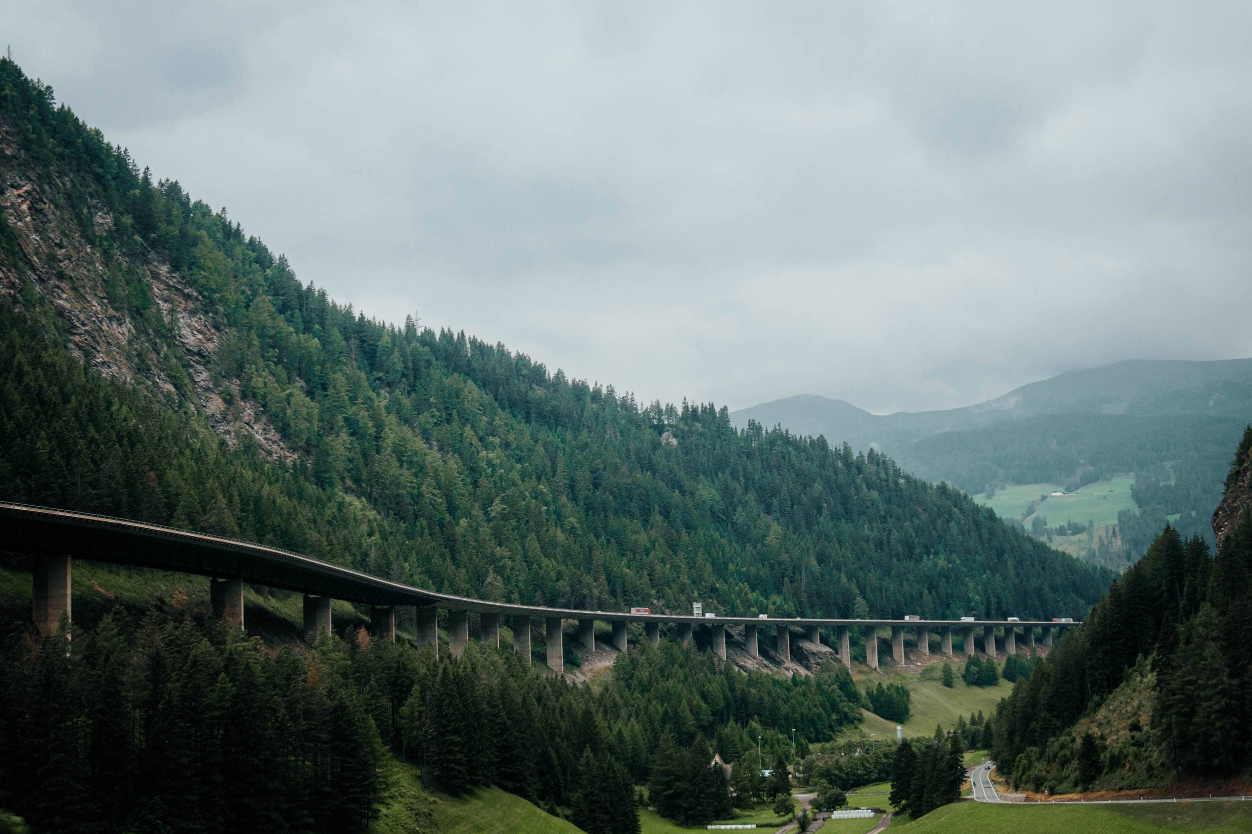 Eine lange Brücke spannt sich durch ein grünes, bewaldetes Tal mit Bergen im Hintergrund unter einem bewölkten Himmel.