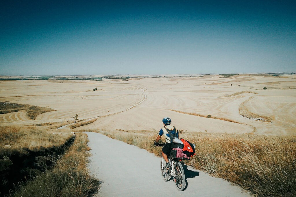 Ein Radfahrer fährt auf einem ländlichen Weg durch weite, goldene Felder unter einem strahlend blauen Himmel.