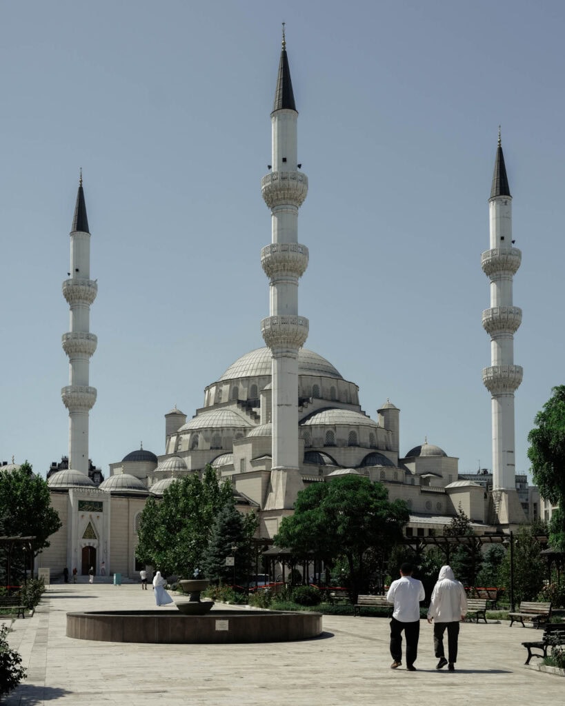 Zwei Menschen gehen auf eine große Moschee mit vier hohen Minaretten zu, die von Bäumen und einem Innenhof umgeben ist.