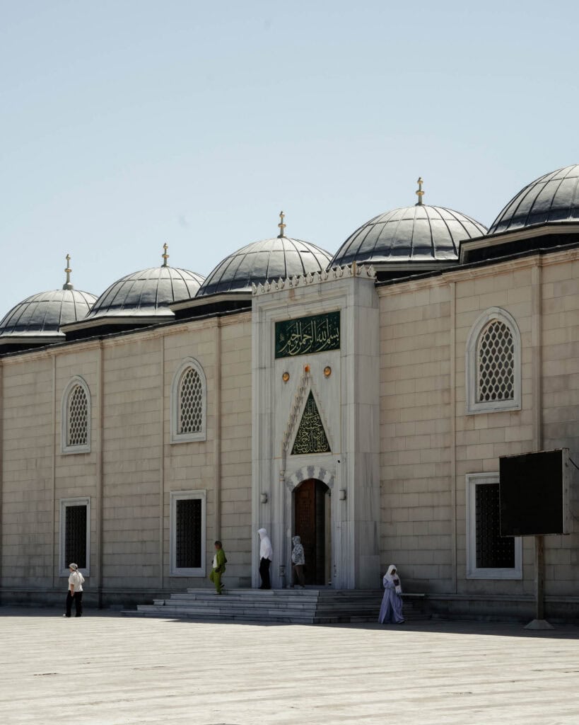 An einem sonnigen Tag gehen Menschen am Eingang einer Moschee mit Kuppeldächern und Bogenfenstern vorbei.