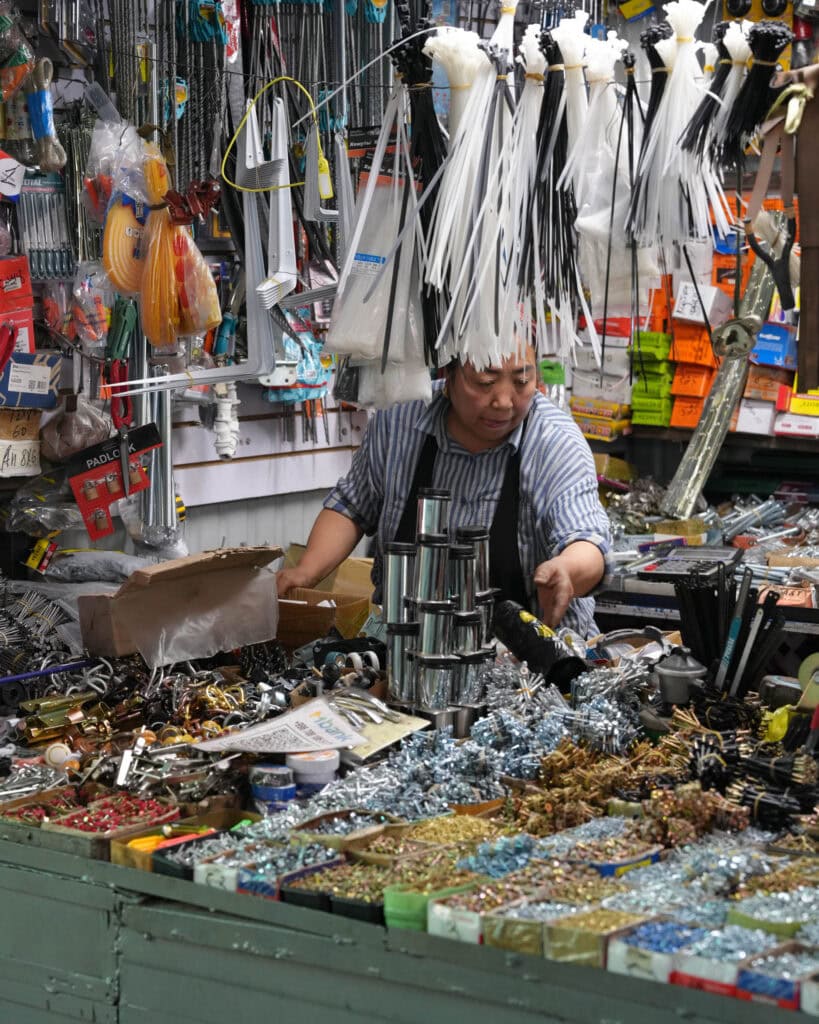 Ein Verkäufer ordnet Waren an einem überfüllten Marktstand an, der mit Werkzeugen, Kleinigkeiten und Bedarfsartikeln gefüllt ist.