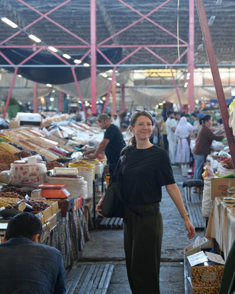 Eine Frau, die lächelnd durch einen belebten Markthallenmarkt geht, in dem sich Verkaufsstände mit Waren befinden und um sie herum Menschen einkaufen.