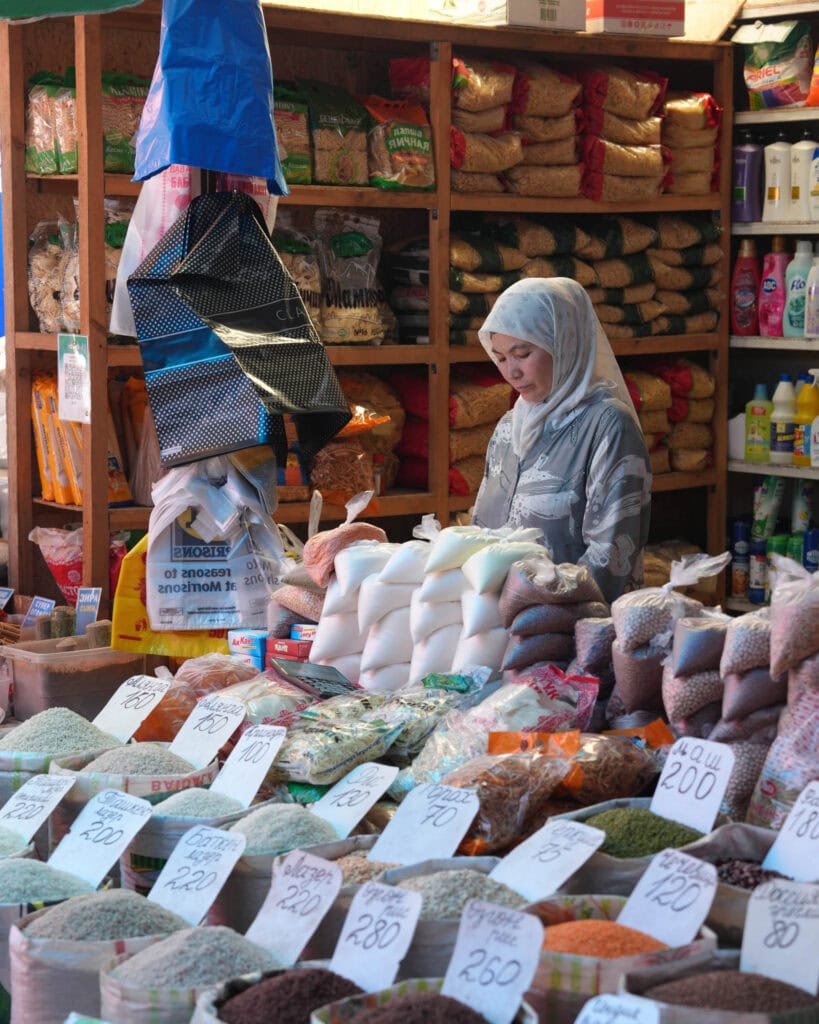 Eine Frau mit Kopftuch verkauft Getreide und Bohnen an einem Marktstand mit handgeschriebenen Preisschildern.