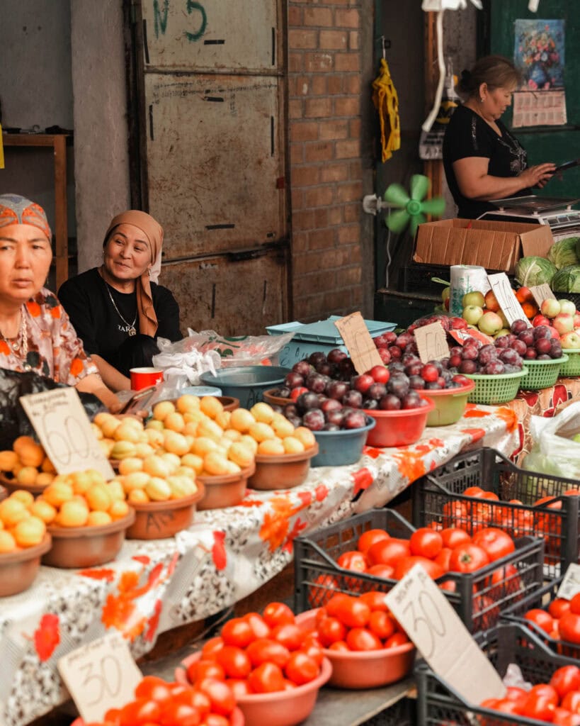 Frauen verkaufen frisches Obst und Gemüse an einem farbenfrohen Marktstand im Freien mit handgeschriebenen Preisschildern.