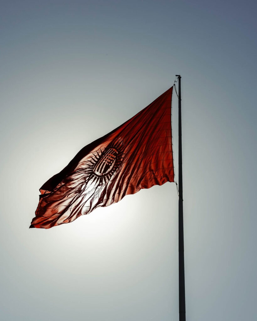 Eine rote Flagge mit einer gelben Sonne und einem Muster, die an einem hohen Fahnenmast vor einem strahlend blauen Himmel weht.