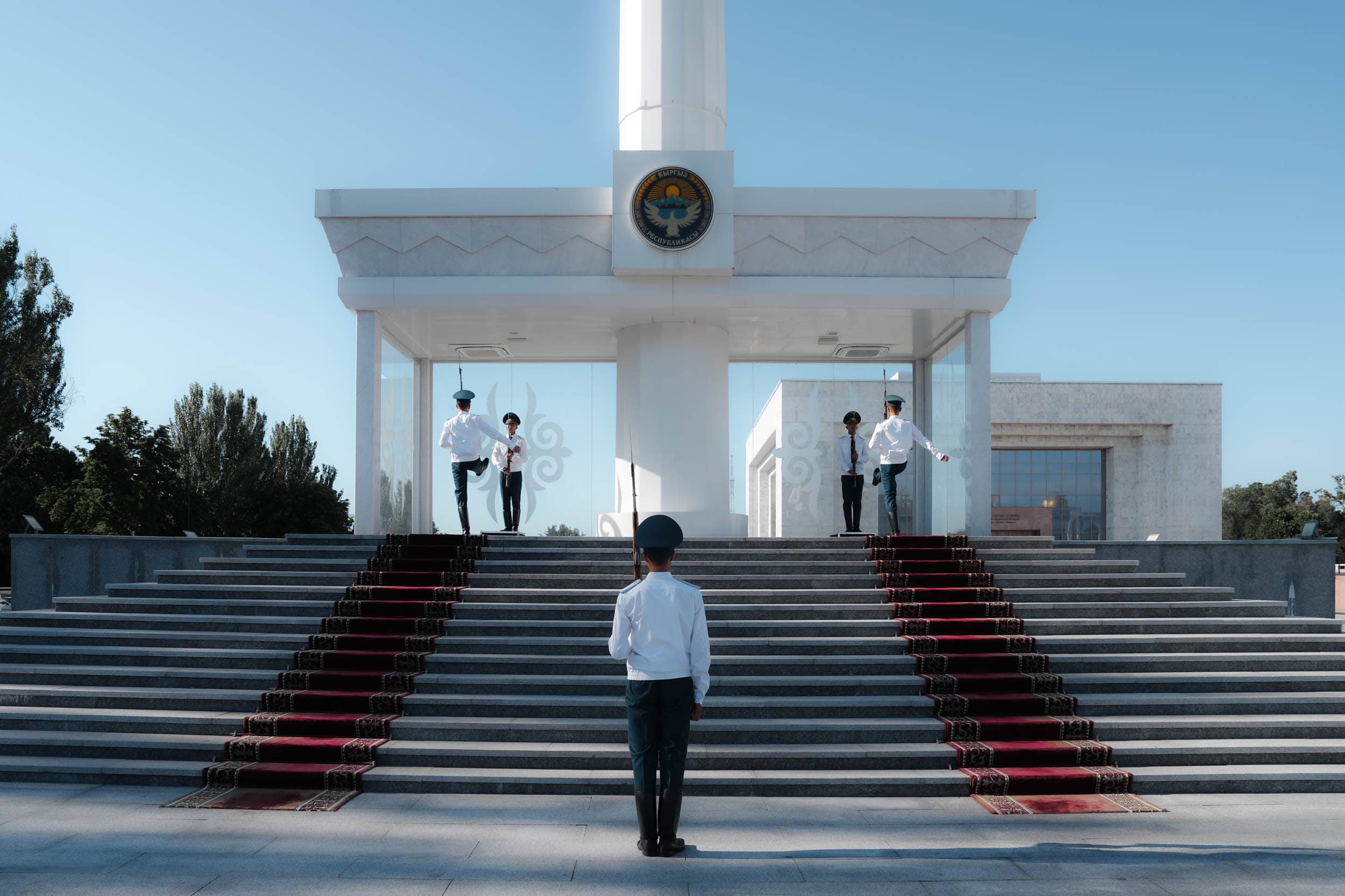 Vier uniformierte Wachen stehen auf einer mit roten Teppichen ausgelegten Treppe und blicken auf ein weißes Denkmal auf einem öffentlichen Platz.