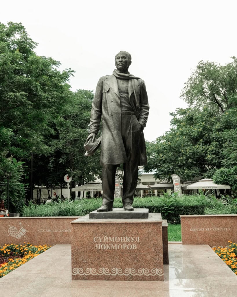 Statue eines Mannes in Mantel und Schal, der einen Hut in der Hand hält und auf einem Sockel in einem Park steht.