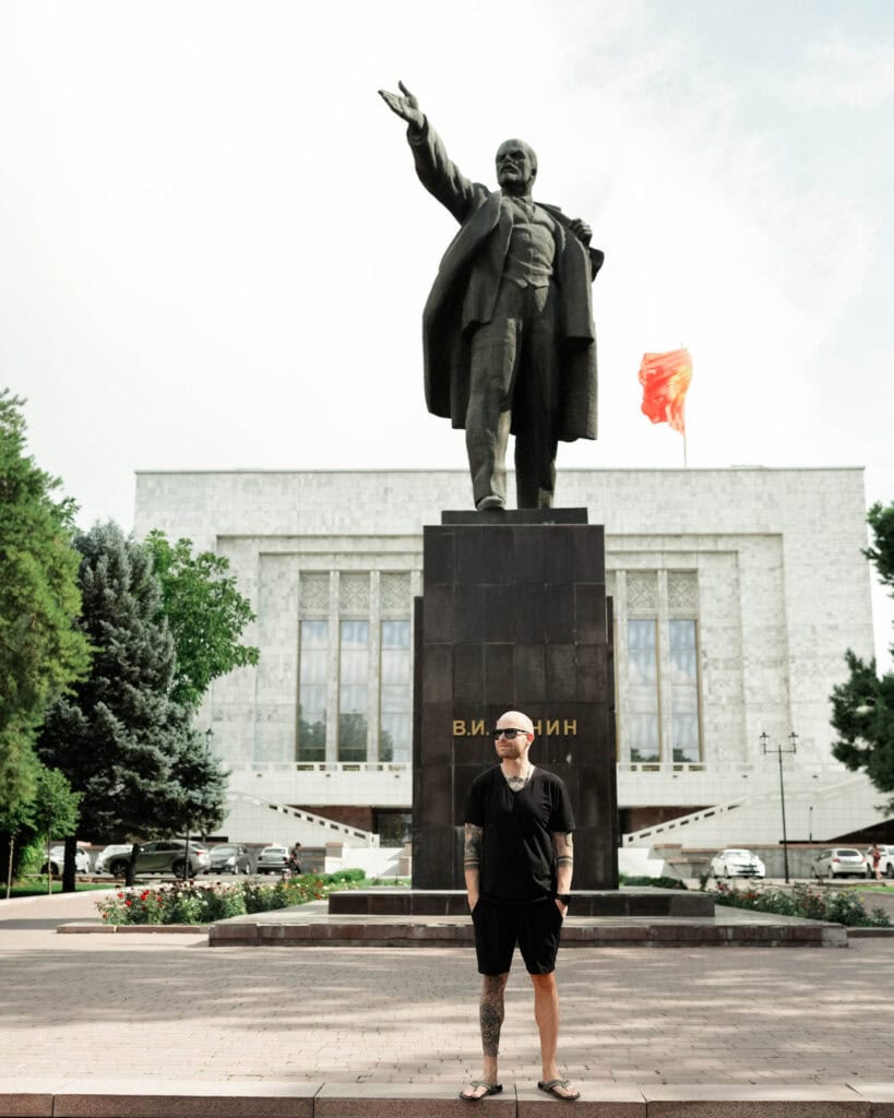 Eine Person in Freizeitkleidung steht vor einer großen Lenin-Statue und einem Gebäude mit einer roten Fahne.