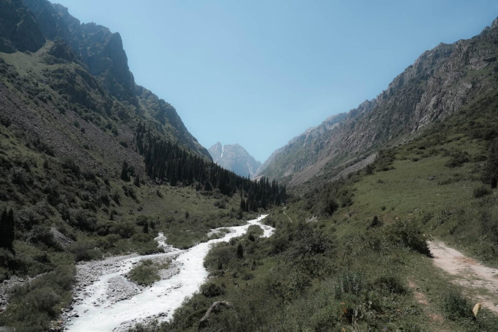 Ein Fluss schlängelt sich unter einem strahlend blauen Himmel durch ein grünes Bergtal, das auf beiden Seiten von Bäumen und schroffen Hügeln gesäumt ist.