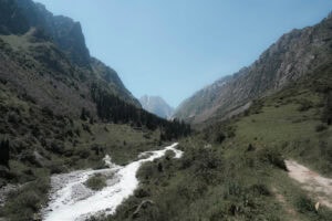 Ein Fluss schlängelt sich unter einem strahlend blauen Himmel durch ein grünes Bergtal, das auf beiden Seiten von Bäumen und schroffen Hügeln gesäumt ist.