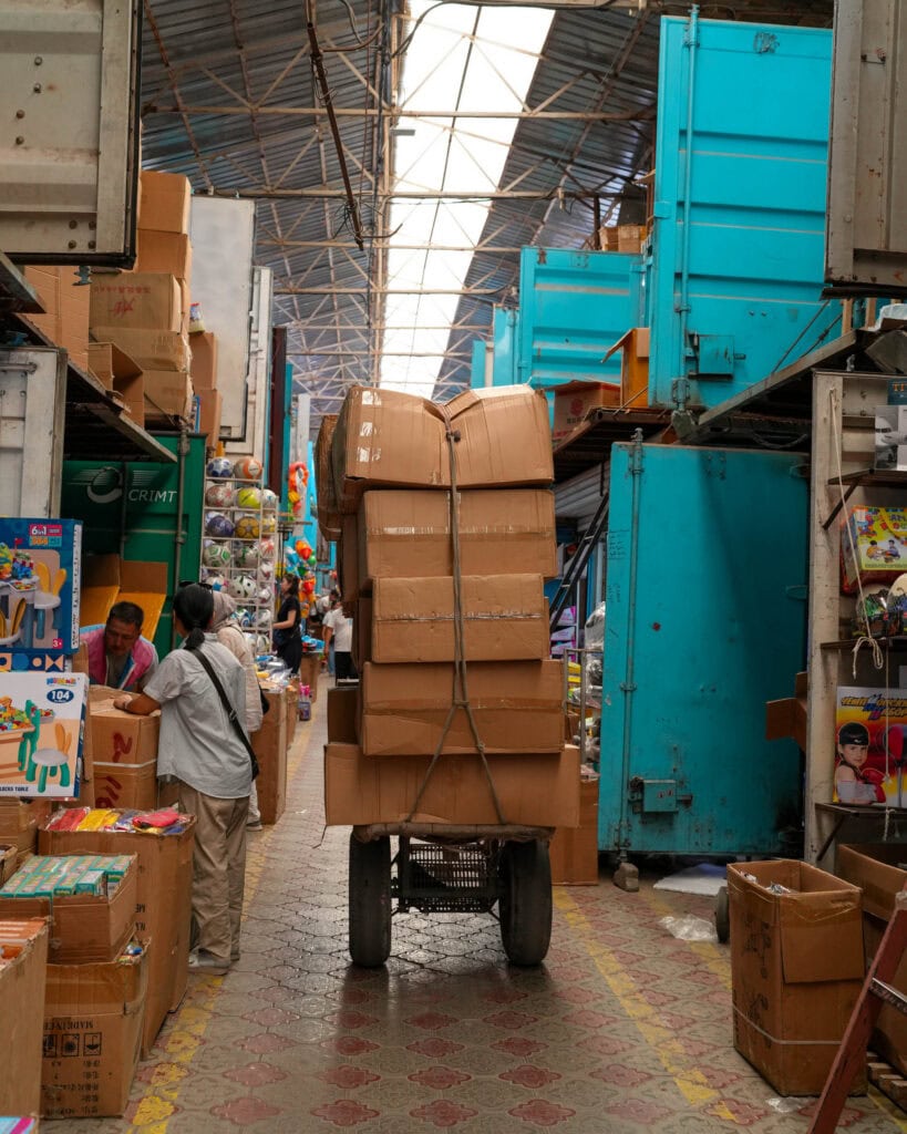 Ein mit Kartons beladener Einkaufswagen fährt durch einen belebten Marktgang, an dessen Seiten sich Geschäfte und Menschen drängen.