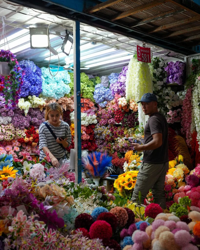 Zwei Menschen kaufen Blumen an einem lebhaften Marktstand, der mit bunten Kunstblumen gefüllt ist.