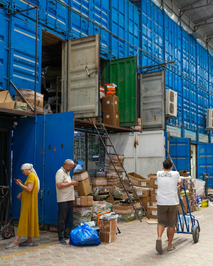 Menschen schauen auf ihre Handys, während hinter ihnen gestapelte Versandcontainer stehen; ein Mann schiebt einen mit Paketen beladenen Wagen auf einem gepflasterten Gehweg.