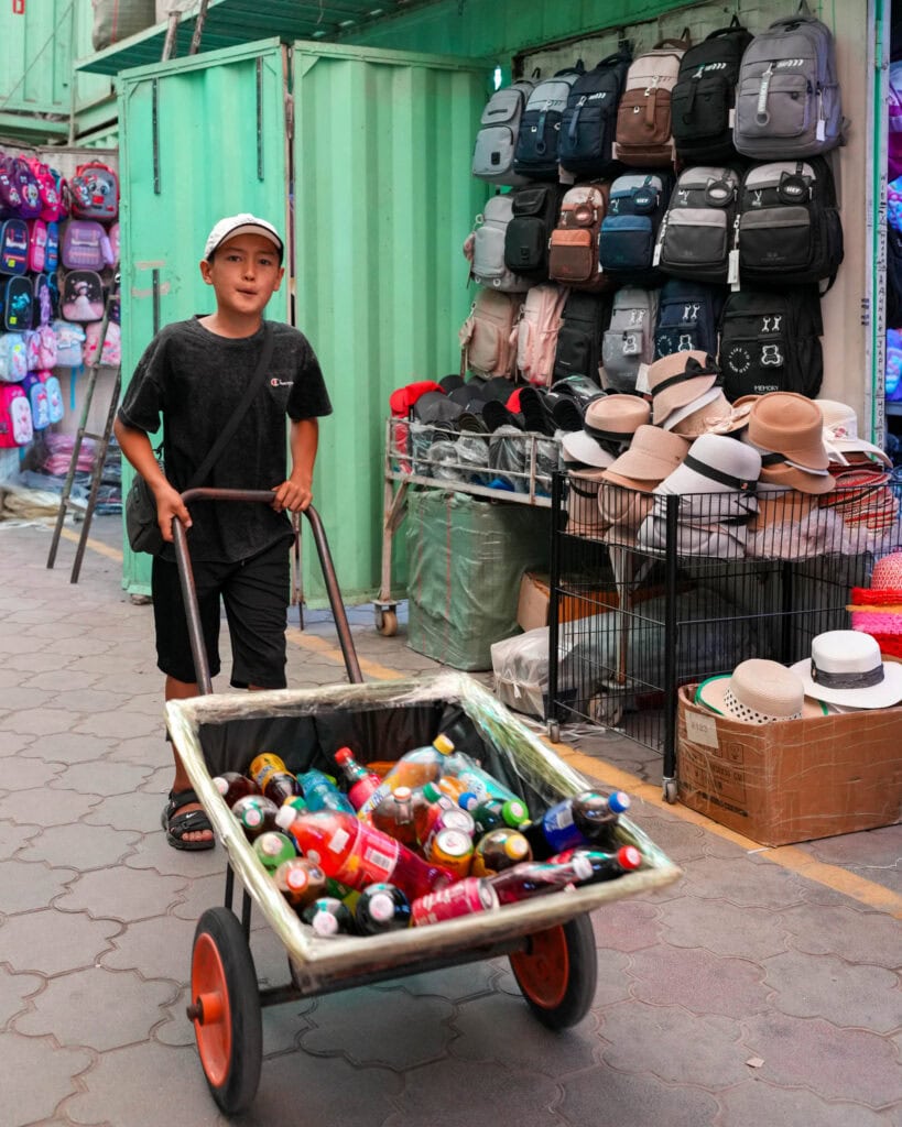 Ein Junge schiebt einen Wagen mit Getränkeflaschen an einem Stand vorbei, an dem Hüte und Rucksäcke verkauft werden, auf einem Markt unter freiem Himmel.